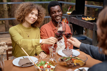 Young happy friends toasting with red wine at table during their meeting at party outdoors