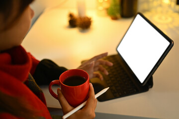 Young woman holding cup of hot coffee and using digital tablet in decorated room with warm light