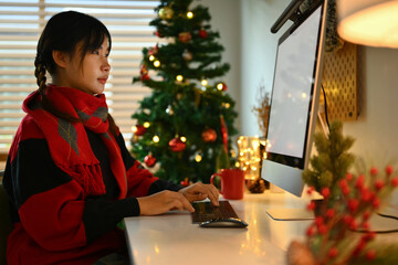 Young woman wearing warm cozy knitted sweater sitting by decorated Christmas tree and using computer