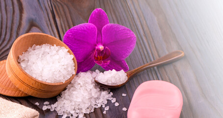 White bath salt in a wooden bowl with a spoon, soap, burlap and orchid on wooden table on sunlight