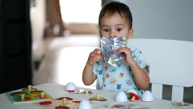 Adorable baby boy sits at desk drinking juice from doypack. Kid looks at the toys scattered in front of him.
