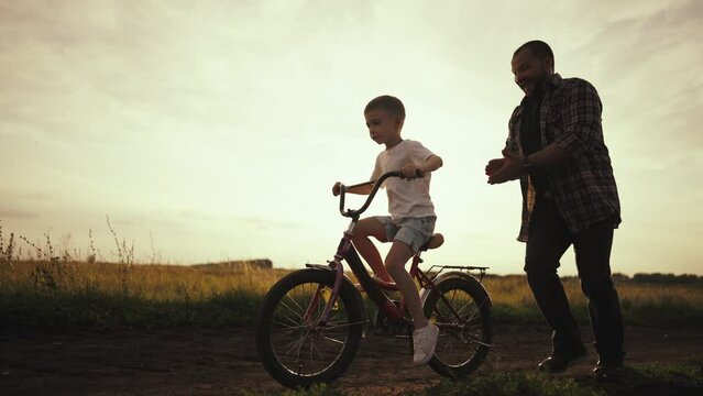 Dad teaches son to ride bike on road. Daddy and kid play together outdoors. Father helps son learn. Child rides bike. Kid riding bicycle while father runs along holding his. Science and sports concept