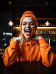 Young, cheerful woman in a bar wearing a orange hoodie and cap