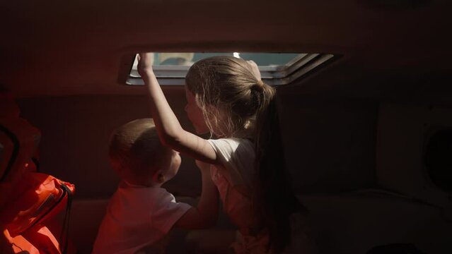 Cheerful children talk and lean out of boat hatch playing in cabin on ocean trip. Cute brother and sister enjoy yacht sailing on tropical vacation. Sea journey