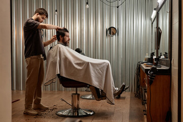Customer sitting in chair and looking at mirror while barber making hairstyle to him in barber shop