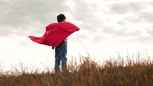 Boy stands in field extending arms wearing red raincoat in overcast weather