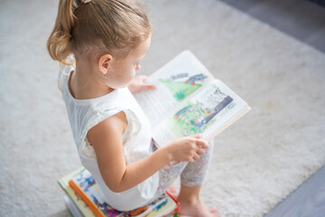 Little girl is sitting on stack of children's books and leafing through a book with pictures of...