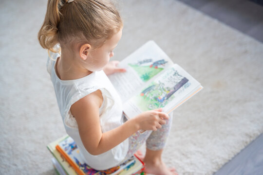 Little girl is sitting on stack of children's books and leafing through a book with pictures of fairy tales