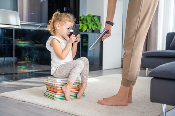 Little girl sits on a stack of children's books and uses her smartphone while mother gives her a...