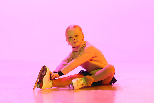 Little adorable baby girl, chide in sweater and skirt, sitting on floor and tying laces on skates against pink background in neon. Concept of childhood, figure skating sport, hobby, school, education