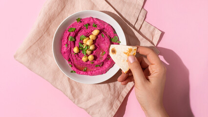 Female hand dipping pita bread triangles in bowl of beetroot hummus on pink background, top view,...