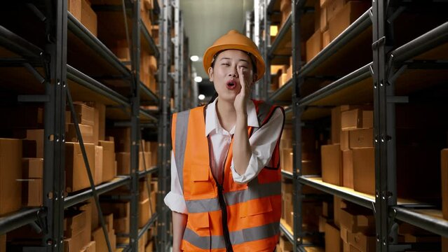 Asian Female Engineer With Safety Helmet Standing In The Warehouse With Shelves Full Of Delivery Goods. Taking Note On The Tablet And Looking Around In The Storage
