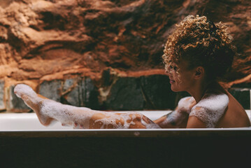 Side view of relaxed african american naked young woman taking bath at home, laying in beautiful bathtub, rubbing her skin with sponge and smiling, copy space. Home spa, hygiene, relaxation concept