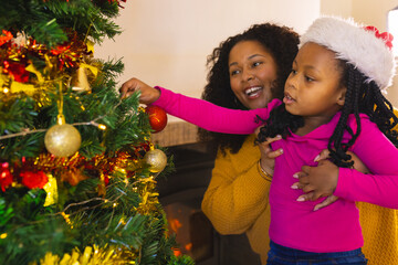 Happy african american mother and daughter decorating christmas tree at home, copy space