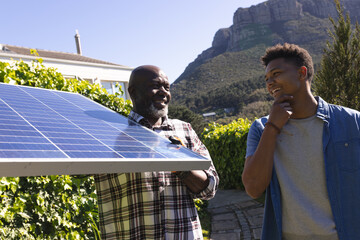 Happy african american father and adult son with solar panel talking in sunny garden