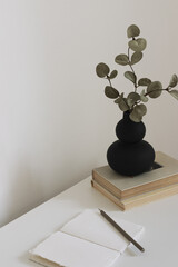 Fototapeta na wymiar Modern black vase with eucalyptus branches, books and a notebook with pencil. Workspace in neatral colors, copy space for your text.
