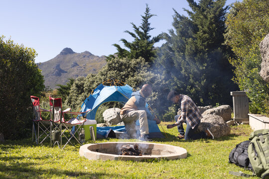 African american father and adult son pitching tent by firepit at sunny campsite, copy space