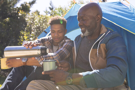 Happy african american father and adult son sitting outside tent having coffee in the sun