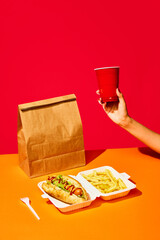 Tasty hotdog with sausage served mustard and ketchup with green salad with refreshing drinks and fried potato against vivid red background. Concept of fast food, delivery.