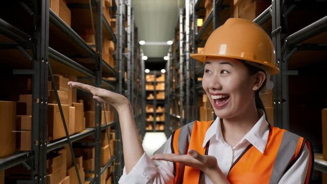 Close Up Of Asian Female Engineer With Safety Helmet Standing In The Warehouse With Shelves Full Of Delivery Goods. Smiling And Pointing To Side Recommends About Something In The Storage
