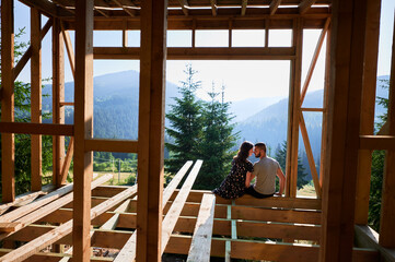 Fototapeta na wymiar Man and woman inspecting their future wooden frame house nestled in the mountains near forest. Youthful couple at construction site in early morning. Concept of contemporary ecological construction.
