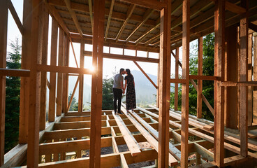 Naklejka na ściany i meble Man and woman inspecting their future wooden frame house nestled in the mountains near forest. Youthful couple at construction site in early morning. Concept of contemporary ecological construction.
