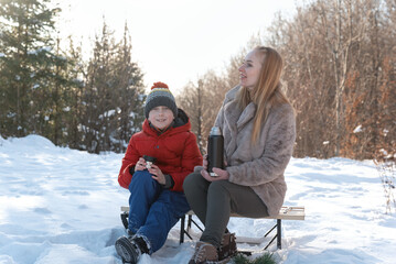 Young woman and boy sit on sled in woods and drink hot tea. Mother and son in snow-covered forest.