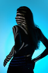 Back view of elegant young woman in jeans, looking away, posing against blue background in neon light. Stripes of shadow. Concept of modern art, beauty, style, futurism and cyberpunk, creativity