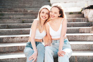 Two young beautiful smiling hipster female in trendy summer white top and jeans clothes. Carefree women posing in the street. Positive models having fun outdoors. Cheerful and happy. Sit at stairs