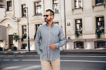 Portrait of handsome confident stylish hipster lambersexual model. Sexy modern man dressed in blue shirt and trousers. Fashion male posing on street background in Europe city at sunset. In sunglasses