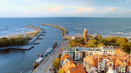 Drone photo captures Kołobrzeg's maritime charm, featuring the iconic lighthouse, cerulean sea, turbulent waves, a distant pier, and autumnal hues on the trees. - obrazy, fototapety, plakaty
