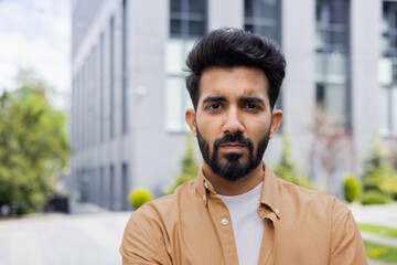 Close-up portrait of young Indian man, serious man concentrating and thoughtfully looking at camera, businessman outside office building. - obrazy, fototapety, plakaty