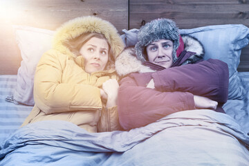 A man and a woman in winter outerwear are freezing under a blanket in bed, the sun's rays are breaking through from above. The concept of cold in the house