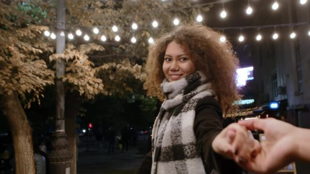 First person view of a young beautiful woman in a coat and scarf leads by the hand and smiles during a joint walk in an evening city street. Concept of cozy and comfortable autumn life