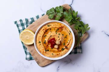 Roasted red pepper hummus in white bowl on white marble background. Top view