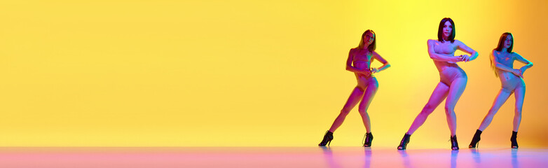 Full-length image of three young women with perfect body shapes dancing on heeled shoes over gradient yellow background in neon light. Concept of hobby, contemporary dance style, art, beauty, elegance