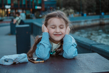 A little girl is sitting near a river in a big city with her eyes and ears closed. A mobile phone...