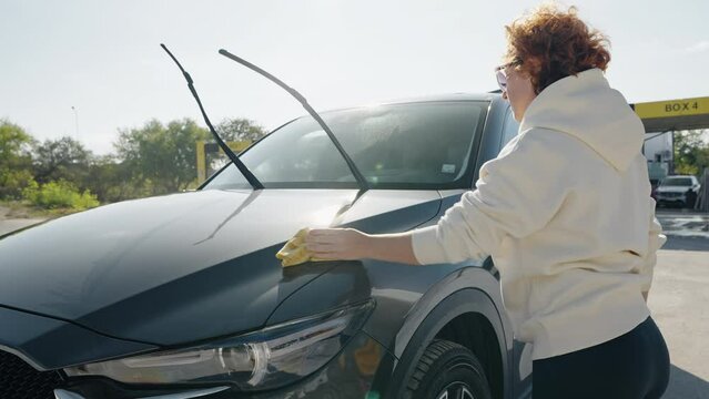 Red-haired woman wipes her car dry from water droplets with rag after washing it wash self-service and sprays foam from hose with pressure washer at car wash station. Lifestyle