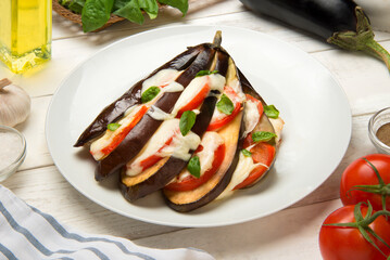 Fan of baked eggplants with tomatoes and mozzarella in a plate on a white rustic table with ingredients.