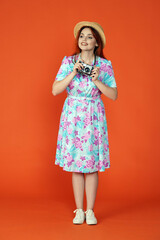 Woman standing holding old film camera, looking at the attractions around and smiling, isolated on orange background. Portrait of female traveler.