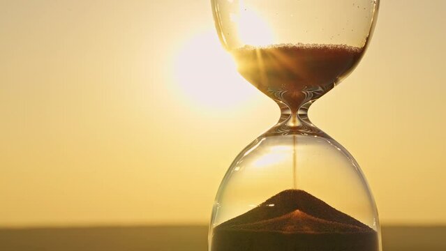 Hourglass concept on blurred background of golden sun disk sky in sunset rays in summer closeup. Golden sand pours quickly. Time concept sand. Relax