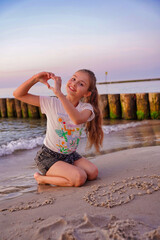 The girl is kneeling on the beach and forming the shape of a heart with her hands.