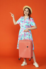 Woman looking at the camera holding suitcase, pointing at the empty area for advertisement, isolated on orange background. Portrait of female traveler.