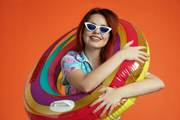 Close up of woman in glasses hugging colorful inflatable pool tube and smiling at the camera, isolated on orange background. Portrait of female traveler.