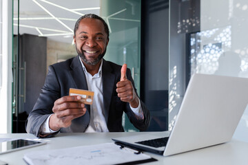 Mature successful businessman smiling and looking at camera, showing thumbs up, holding bank credit...