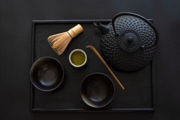 Aerial view of black tray with traditional oriental teapot and bowls, whisk and wooden spoon, matcha tea, black background, horizontal