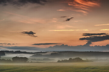 Beautiful sunrise with mist against a dramatic and colorful sky background. Calm and breathtaking landscape view on a field in the countryside with blooming trees on a foggy and overcast morning