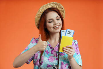 Close up of woman in straw hat holding flight tickets, passport looking at them and showing thumbs up, isolated on orange background. Portrait of female traveler.