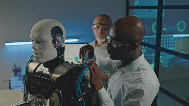 Waist up of Black male engineer working with screwdriver while repairing humanoid robot in workshop and talking to colleague checking operating system on digital tablet