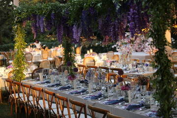 Decorated white wedding table for a festive dinner with pink flowers in brass pots on green lawn under the open sky.